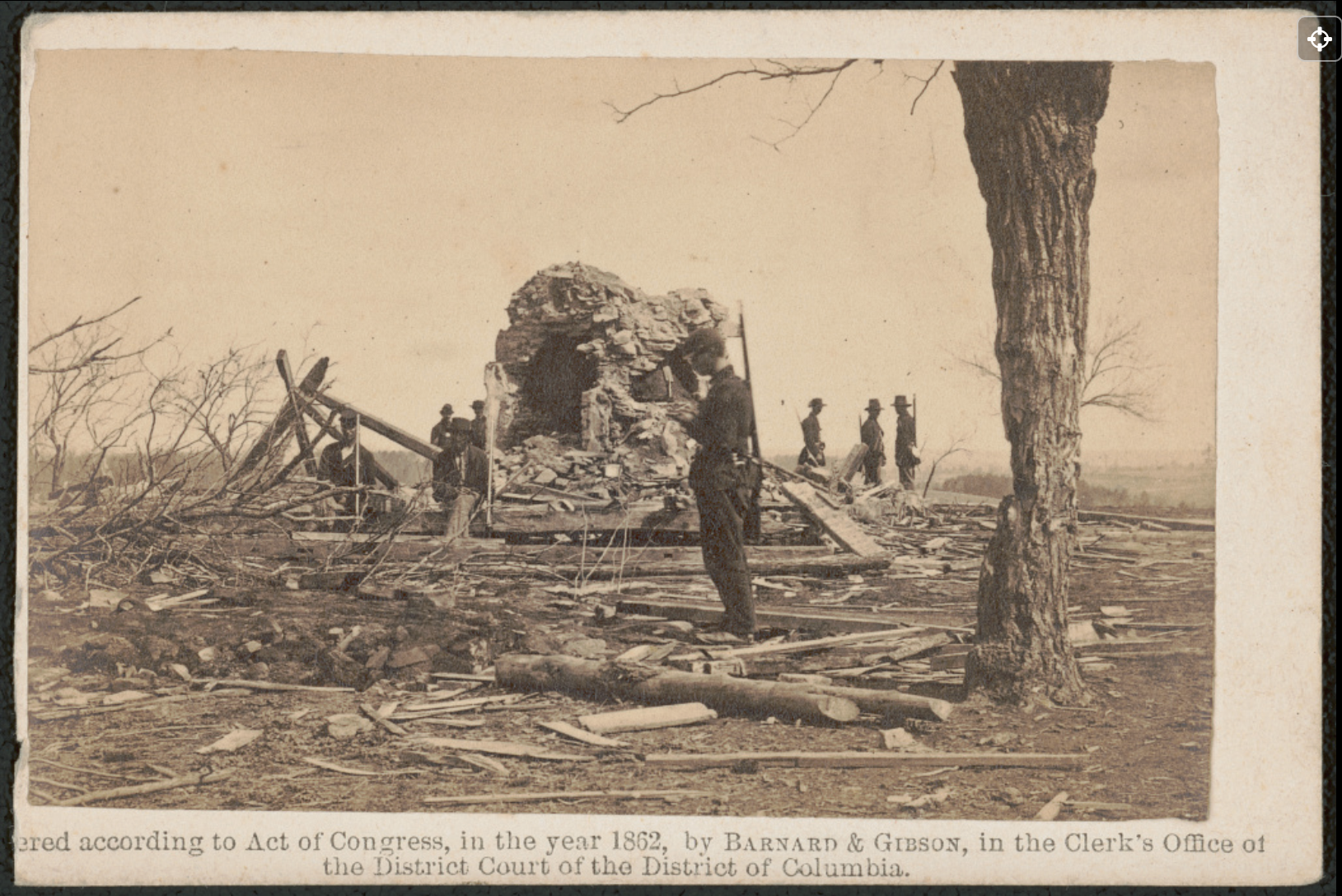 Sepia-colored photo of stone ruins of a house among destroyed trees, with men standing around.