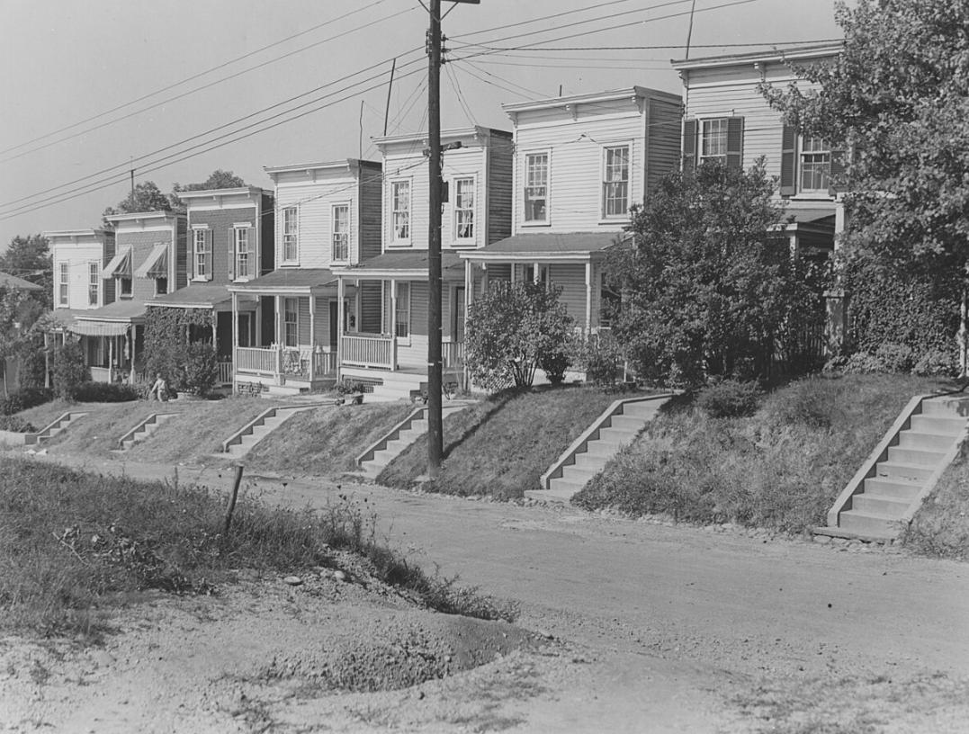 A black-and-white image of a street with 7 row houses on a hill. (Source: National Park Service Museum Resource Center, Public domain, via Wikimedia Commons)