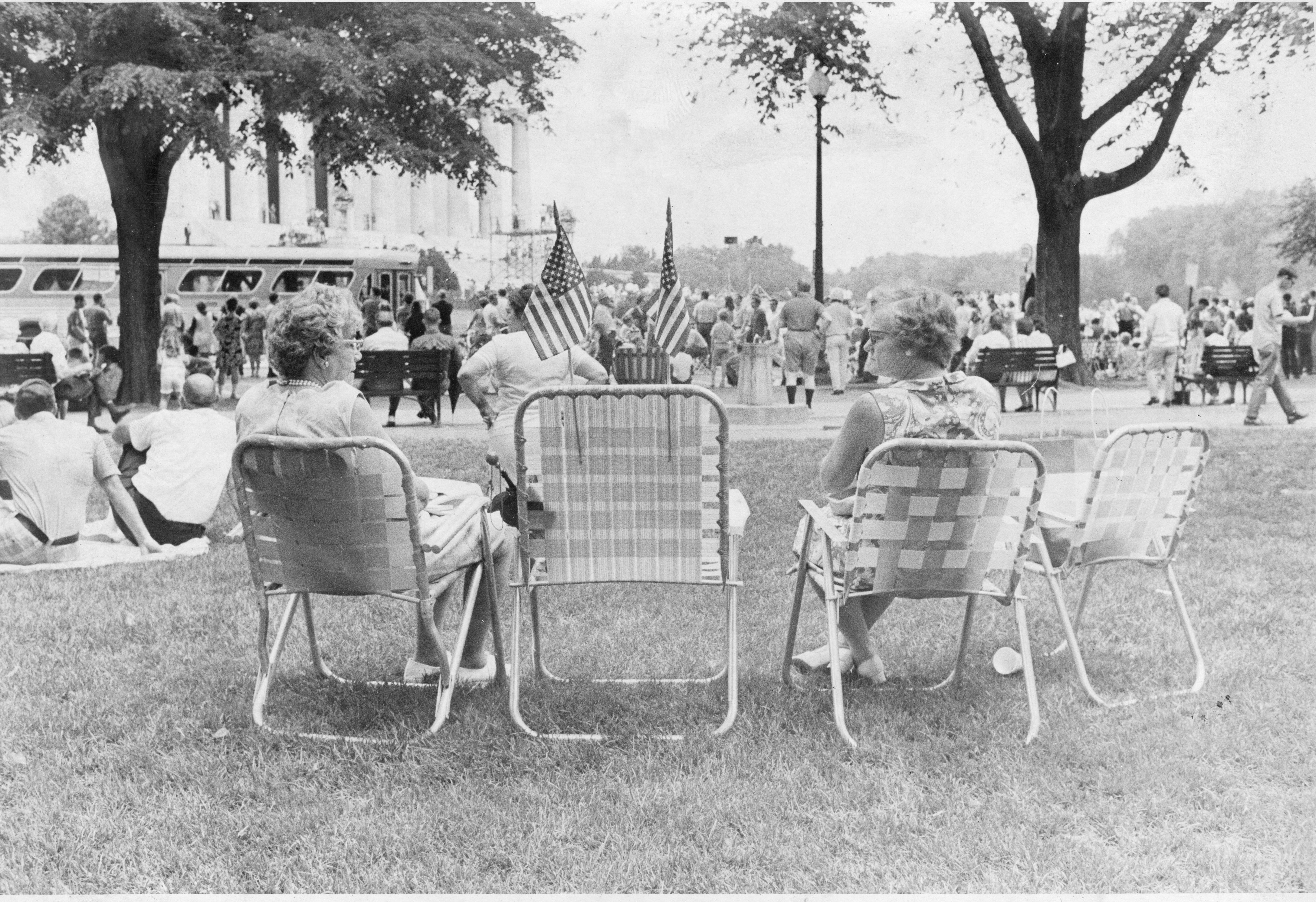 Three older, white spectators seat in lawnchairs watching Honor America Day programming by the Lincoln Memorial.