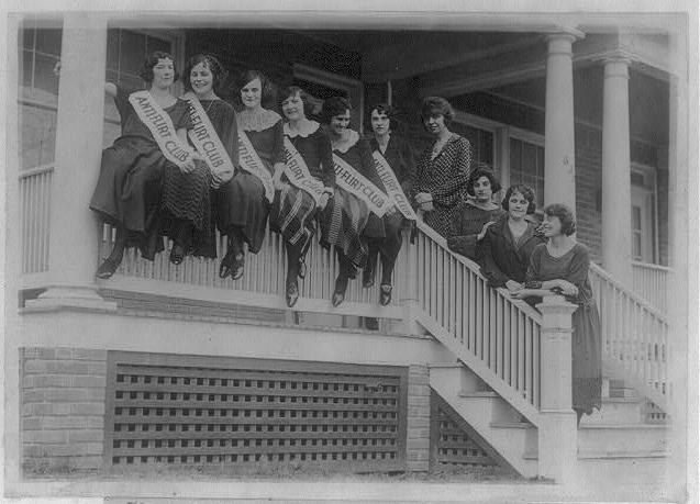 A group of women sit on the railing of a porch wearing banners across their chests that read "Anti-flirt club"