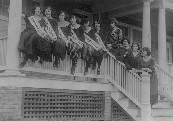 A group of women sit on the railing of a porch wearing banners across their chests that read "Anti-flirt club"