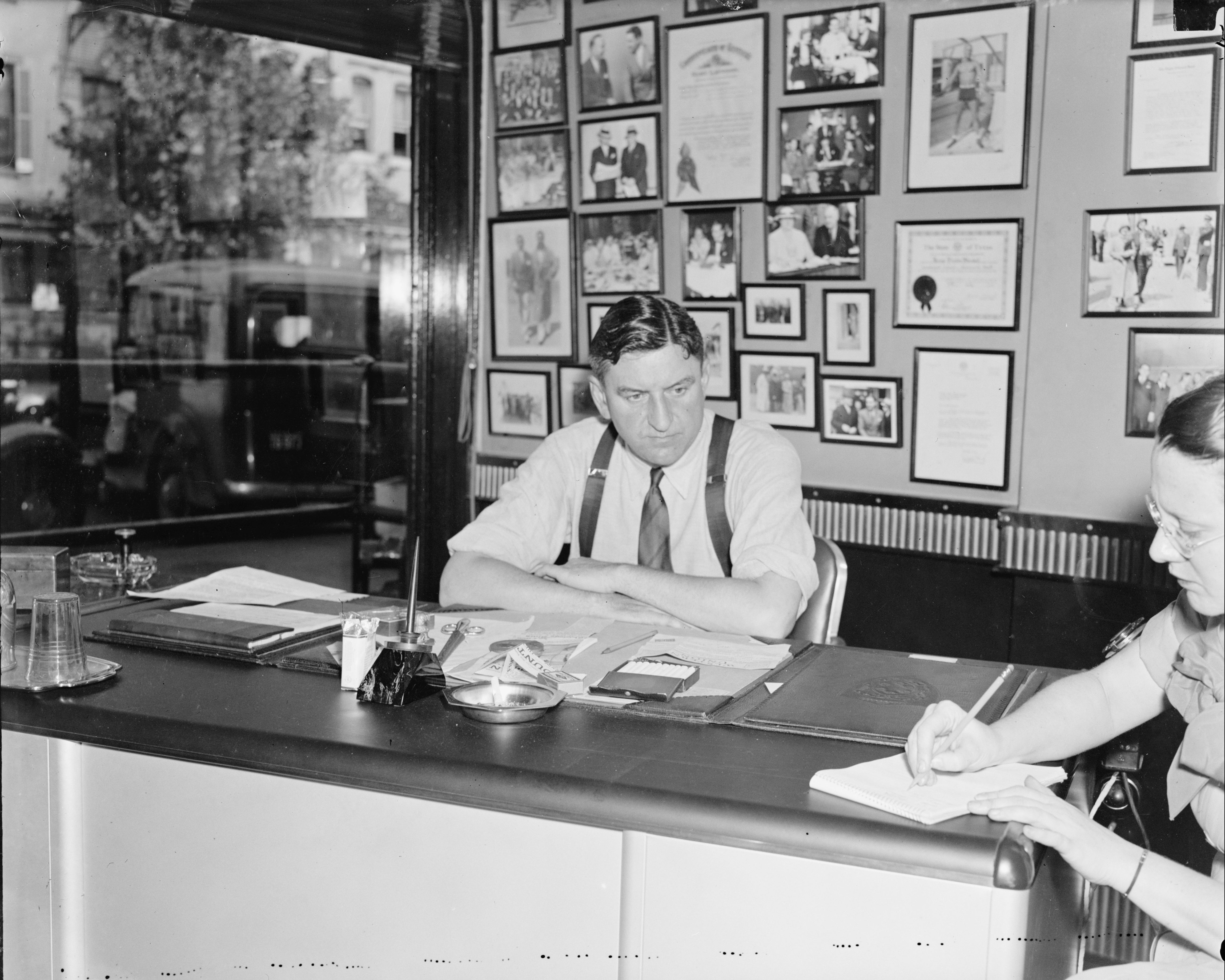 Business man wearing tie sits at a desk talking to a woman at far right of frame, who is taking notes.