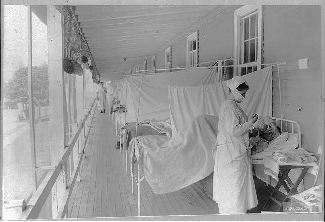 A black and white photograph of a nurse caring for a line of men stricken with the Spanish Flu at Walter Reed Hospital. They are on a covered porch, the Nurse wears  1910s personal protective equipment. 