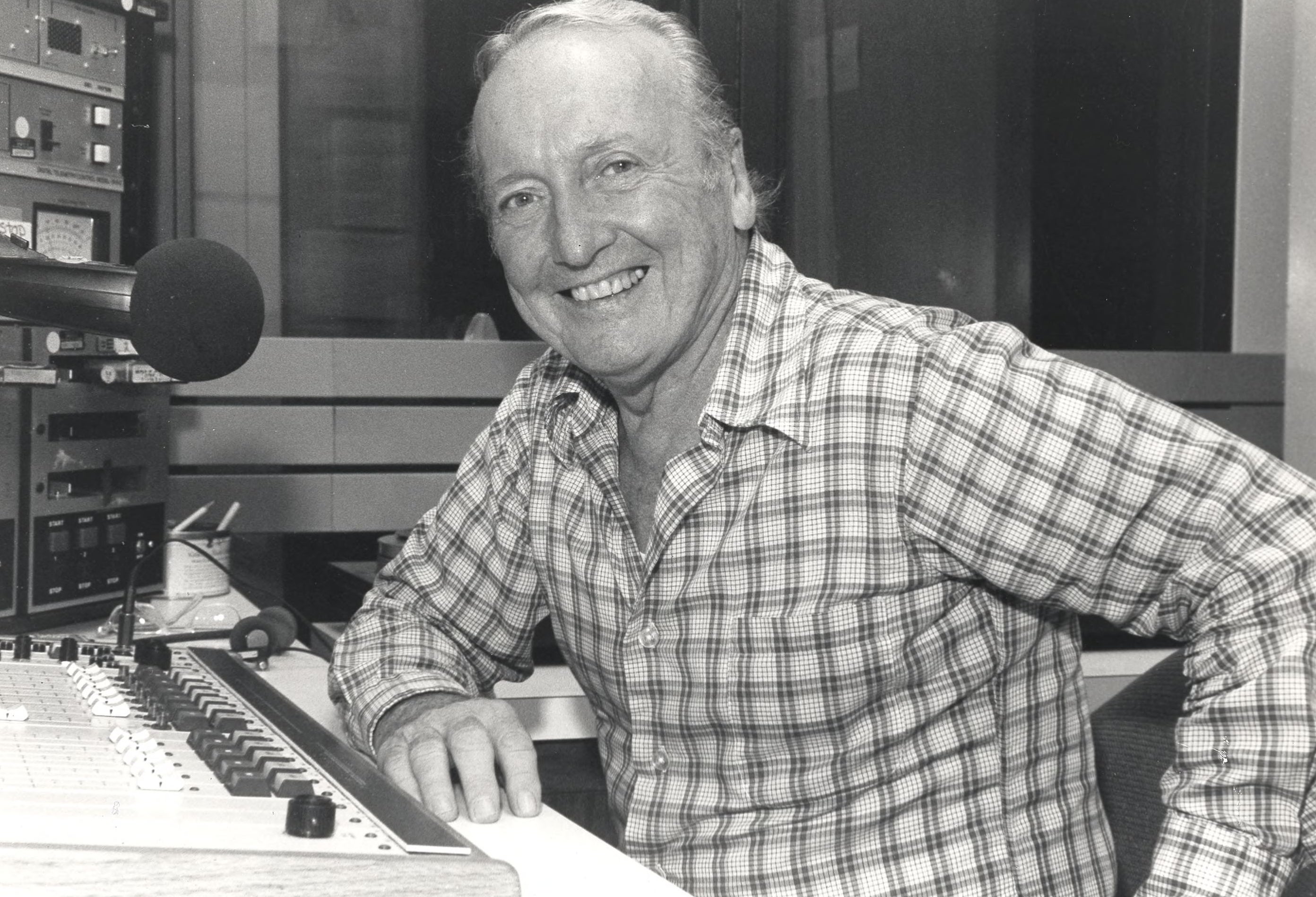 An older Grant sits at a desk in a radio station booth. He has an elbow on the table and gives a big smile to the camera.