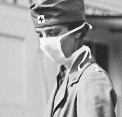 This is a black and white photograph of a woman wearing a mask, she wears a hat with the familiar plus sign associated with the Red Cross. Despite the mask covering most of her face, she wears a serious expression. 