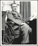 A black-and-white photo. Hugo Deffner, a white man in his 60s, sits in his wheelchair at a desk. He is holding a piece of paper in one hand and wearing a suit. He is looking at the camera.